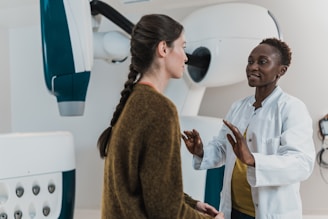 Two women are engaged in conversation in a medical setting, one wearing a white coat, suggesting she is a healthcare professional. The environment contains medical equipment, possibly indicating a check-up or consultation is taking place.
