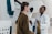 Two women are engaged in conversation in a medical setting, one wearing a white coat, suggesting she is a healthcare professional. The environment contains medical equipment, possibly indicating a check-up or consultation is taking place.