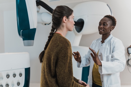 Two women are engaged in conversation in a medical setting, one wearing a white coat, suggesting she is a healthcare professional. The environment contains medical equipment, possibly indicating a check-up or consultation is taking place.