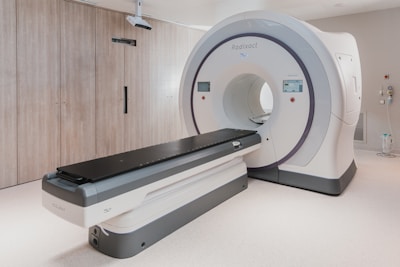 A large, modern medical imaging machine, possibly a CT scanner, is situated in a clean and well-lit medical room. The machine is white with grey accents and features a circular opening with a long table extending from it, which can slide the patient in for scanning. On the side of the scanner, there are control panels with digital displays and buttons.