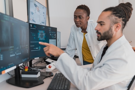 Two people are working in a lab environment, both wearing lab coats. One person is seated and pointing at a computer screen, while the other is standing, looking at the screens. Multiple computer monitors display complex data, possibly related to scientific or medical research.