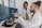 Two people are working in a lab environment, both wearing lab coats. One person is seated and pointing at a computer screen, while the other is standing, looking at the screens. Multiple computer monitors display complex data, possibly related to scientific or medical research.