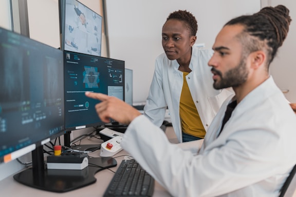 Two people are working in a lab environment, both wearing lab coats. One person is seated and pointing at a computer screen, while the other is standing, looking at the screens. Multiple computer monitors display complex data, possibly related to scientific or medical research.