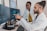 Two people are working in a lab environment, both wearing lab coats. One person is seated and pointing at a computer screen, while the other is standing, looking at the screens. Multiple computer monitors display complex data, possibly related to scientific or medical research.