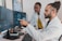 Two people are working in a lab environment, both wearing lab coats. One person is seated and pointing at a computer screen, while the other is standing, looking at the screens. Multiple computer monitors display complex data, possibly related to scientific or medical research.