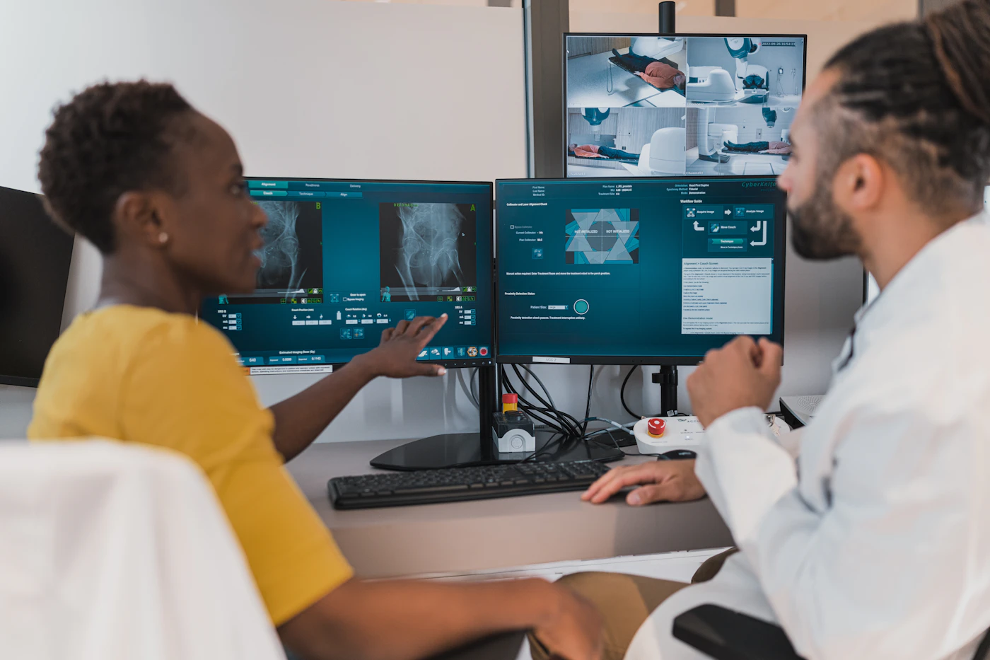 A clinician in a white coat reviewing health data on a desktop screen while talking with a colleague in a bright clinic office