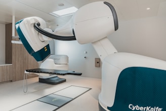 A modern medical device with large mechanical arms, likely used for precise medical procedures. It is situated in a clinical environment with clean, white walls and a tidy appearance. The equipment is labeled 'CyberKnife', indicating its purpose in advanced medical treatments.