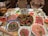 A table is set with an assortment of food items for a hot pot meal. In the center, there is a pot with divided sections containing various broths and chilies. Surrounding the pot are plates of raw meat slices, vegetables, and condiments. There are bowls of watermelon slices and leafy greens. A person is pouring a drink from a pitcher on the left side of the table.