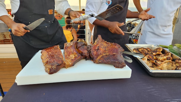 Three cooks are preparing and serving large cuts of roasted meat on a white cutting board. Nearby, a tray filled with grilled pieces of meat rests on the table. They are standing outside with people in the background.
