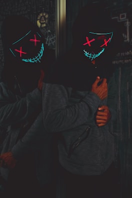 A person wearing a dark hoodie with a glowing, abstract mask featuring bright neon colors, standing in front of a chalkboard. The mask has a sinister smile and crosses for eyes, glowing in red and blue hues. The atmosphere is dark and mysterious.