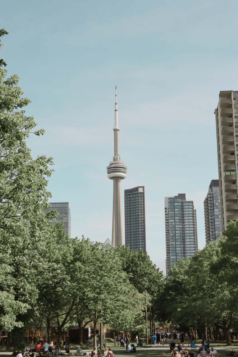 The image depicts a bustling city scene with a tall, white tower dominating the skyline. The tower is part of the CN Tower, which stands out against the backdrop of modern skyscrapers and trees. The perspective of the photo is from ground level, looking up at the tower, giving it an imposing presence. In the foreground, there are people walking around in front of a park with lush green trees, providing a contrast to the concrete structures behind them.