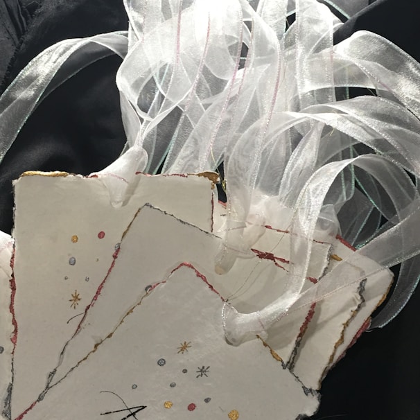 A collection of decorative paper cards with a rough, textured edge, embellished with delicate, colorful designs, and adorned with sheer, white ribbons against a dark background.