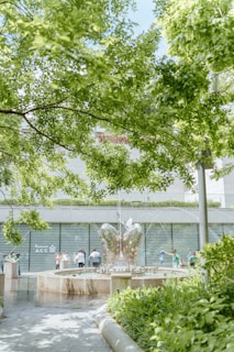 A lush green park features a large, mirrored sculpture shaped like a butterfly with water fountains sprouting from its center. Several people stand around the fountain, engaging with the artwork and enjoying the serene environment. The background showcases a modern building, partially obscured by trees, adding an urban touch to the natural setting.