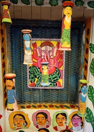 A vibrant and colorful wall art featuring multiple painted clay figures and a central painting of a deity with multiple arms in a seated position. The figures and the deity are painted in bright colors with intricate patterns, and the surrounding walls are adorned with portraits and floral designs.