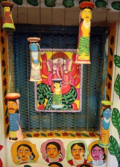 A vibrant and colorful wall art featuring multiple painted clay figures and a central painting of a deity with multiple arms in a seated position. The figures and the deity are painted in bright colors with intricate patterns, and the surrounding walls are adorned with portraits and floral designs.