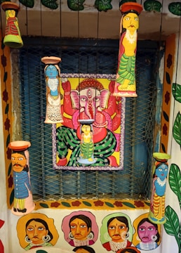 A vibrant and colorful wall art featuring multiple painted clay figures and a central painting of a deity with multiple arms in a seated position. The figures and the deity are painted in bright colors with intricate patterns, and the surrounding walls are adorned with portraits and floral designs.