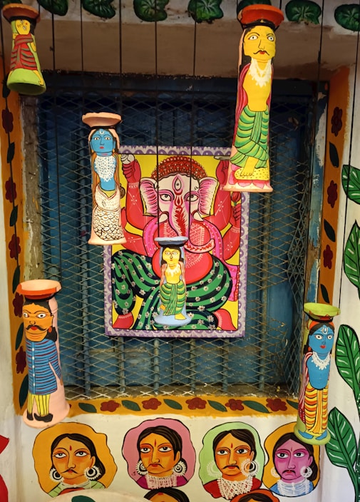 A vibrant and colorful wall art featuring multiple painted clay figures and a central painting of a deity with multiple arms in a seated position. The figures and the deity are painted in bright colors with intricate patterns, and the surrounding walls are adorned with portraits and floral designs.