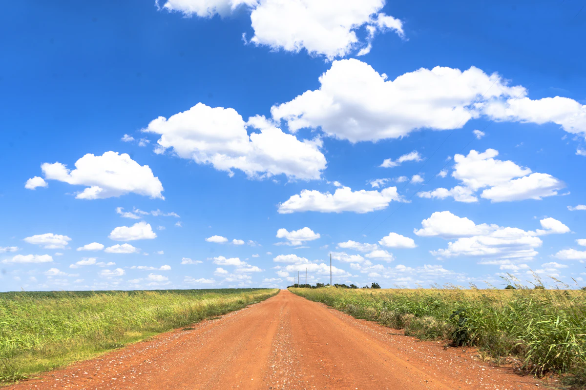 A straight red dirt road between grassy fields under a blue sky with white clouds, near Oklahoma City