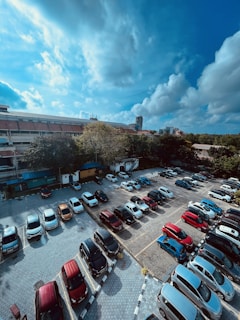 Aerial view of a parking lot filled with various cars. The scene includes red, white, black, and blue vehicles neatly parked in designated spots. In the background, a building with a red-tiled roof is visible, surrounded by lush green trees. The sky above is partially cloudy with patches of blue and white clouds.