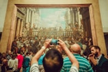 A large, detailed painting is on display in a gallery setting with numerous people gathered around. The artwork features a historical or religious scene with many figures involved in a banquet or gathering. A person in the foreground is taking a photo of the painting with a smartphone.