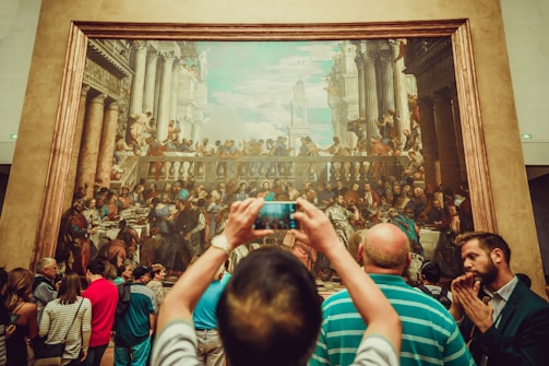 A large, detailed painting is on display in a gallery setting with numerous people gathered around. The artwork features a historical or religious scene with many figures involved in a banquet or gathering. A person in the foreground is taking a photo of the painting with a smartphone.