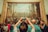 A large, detailed painting is on display in a gallery setting with numerous people gathered around. The artwork features a historical or religious scene with many figures involved in a banquet or gathering. A person in the foreground is taking a photo of the painting with a smartphone.