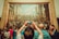 A large, detailed painting is on display in a gallery setting with numerous people gathered around. The artwork features a historical or religious scene with many figures involved in a banquet or gathering. A person in the foreground is taking a photo of the painting with a smartphone.