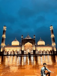 An illuminated mosque stands majestically against a deep blue evening sky. The building features large domes and tall minarets, with the mosque's lights casting a warm glow and reflections on the wet courtyard tiles. A person in a blue shirt is crouching in the foreground.