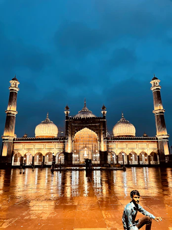 An illuminated mosque stands majestically against a deep blue evening sky. The building features large domes and tall minarets, with the mosque's lights casting a warm glow and reflections on the wet courtyard tiles. A person in a blue shirt is crouching in the foreground.