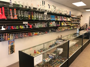 A store interior with shelves and glass display cases filled with a variety of smoking accessories, including glass pipes, bongs, and other paraphernalia. The shelves are well-organized and colorful, showing a range of designs. The space is brightly lit with overhead lights, and the products are arranged in an aesthetically pleasing manner.