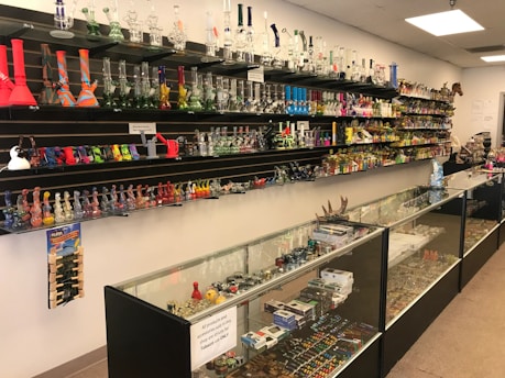 A store interior with shelves and glass display cases filled with a variety of smoking accessories, including glass pipes, bongs, and other paraphernalia. The shelves are well-organized and colorful, showing a range of designs. The space is brightly lit with overhead lights, and the products are arranged in an aesthetically pleasing manner.