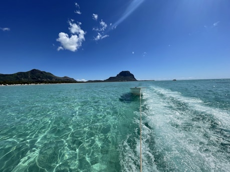 A small boat is tied to a rope, floating on crystal clear turquoise water with a trail of white foam behind it. A mountainous island is visible in the background under a bright blue sky adorned with a few fluffy clouds.