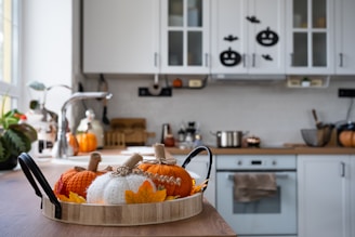 A cozy kitchen decorated for autumn with knitted pumpkins and leaf accents on a wooden tray. The space features a wooden countertop, a stainless steel faucet, and white cabinets adorned with Halloween-themed stickers. A pot sits on the stove, and various kitchen utensils are organized neatly.