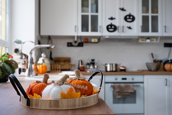 A cozy kitchen decorated for autumn with knitted pumpkins and leaf accents on a wooden tray. The space features a wooden countertop, a stainless steel faucet, and white cabinets adorned with Halloween-themed stickers. A pot sits on the stove, and various kitchen utensils are organized neatly.