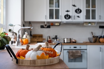 A cozy kitchen decorated for autumn with knitted pumpkins and leaf accents on a wooden tray. The space features a wooden countertop, a stainless steel faucet, and white cabinets adorned with Halloween-themed stickers. A pot sits on the stove, and various kitchen utensils are organized neatly.