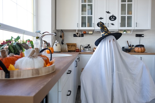 A kitchen with white cabinets and countertops decorated for Halloween. On the right, there is a figure dressed as a ghost with a white sheet and a black witch hat. The left side of the kitchen counter has autumnal decorations including knitted pumpkins and orange leaves. Various kitchen utensils, appliances, and a large pumpkin can also be seen on the countertops.