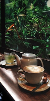 A cozy café scene featuring a table set with a hot latte with foam art, a teapot, and a cup of herbal tea. The background reveals a lush, verdant garden through a large window, allowing sunlight to softly illuminate the setting.