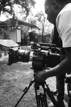 A person operating a professional video camera on a tripod is captured in a monochrome setting. The camera is a high-end model and appears to be set up outdoors, with a blurred background of trees and a building.