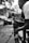 A person operating a professional video camera on a tripod is captured in a monochrome setting. The camera is a high-end model and appears to be set up outdoors, with a blurred background of trees and a building.