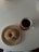 A sugar-coated donut with a bite taken out is placed on a beige plate. Next to the plate is a cup of dark coffee on a wooden coaster, arranged on a round, white table.