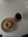 A sugar-coated donut with a bite taken out is placed on a beige plate. Next to the plate is a cup of dark coffee on a wooden coaster, arranged on a round, white table.