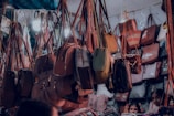 A variety of handbags are hanging in a crowded market stall, each suspended by straps. The bags display different styles, colors, and sizes, creating a vibrant and busy scene. There are people partially visible at the bottom, potentially browsing the collection.