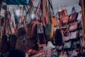 A variety of handbags are hanging in a crowded market stall, each suspended by straps. The bags display different styles, colors, and sizes, creating a vibrant and busy scene. There are people partially visible at the bottom, potentially browsing the collection.