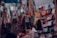 A variety of handbags are hanging in a crowded market stall, each suspended by straps. The bags display different styles, colors, and sizes, creating a vibrant and busy scene. There are people partially visible at the bottom, potentially browsing the collection.