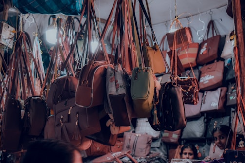 A variety of handbags are hanging in a crowded market stall, each suspended by straps. The bags display different styles, colors, and sizes, creating a vibrant and busy scene. There are people partially visible at the bottom, potentially browsing the collection.