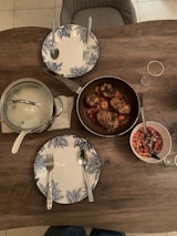 A wooden dining table set for two with blue and white floral patterned plates and silver cutlery. A pot with a glass lid and a pan containing cooked chicken pieces rest on the table. There is also a bowl of chopped vegetables and two empty glasses.