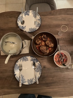 A wooden dining table set for two with blue and white floral patterned plates and silver cutlery. A pot with a glass lid and a pan containing cooked chicken pieces rest on the table. There is also a bowl of chopped vegetables and two empty glasses.