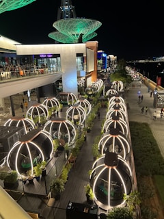 A nighttime scene of a modern, outdoor dining area with glowing dome-shaped seating pods. The setting features an elevated walkway bustling with people and illuminated by vibrant lights. Decorative green structures rise above the scene, adding a futuristic element.