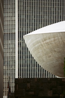 A modern architectural scene featuring a distinctive, large, curved structure with a textured surface, set against a backdrop of vertical lines from a tall building. A stone wall is visible in the foreground, and a small portion of a flag can be seen below.