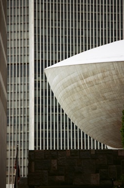 A modern architectural scene featuring a distinctive, large, curved structure with a textured surface, set against a backdrop of vertical lines from a tall building. A stone wall is visible in the foreground, and a small portion of a flag can be seen below.